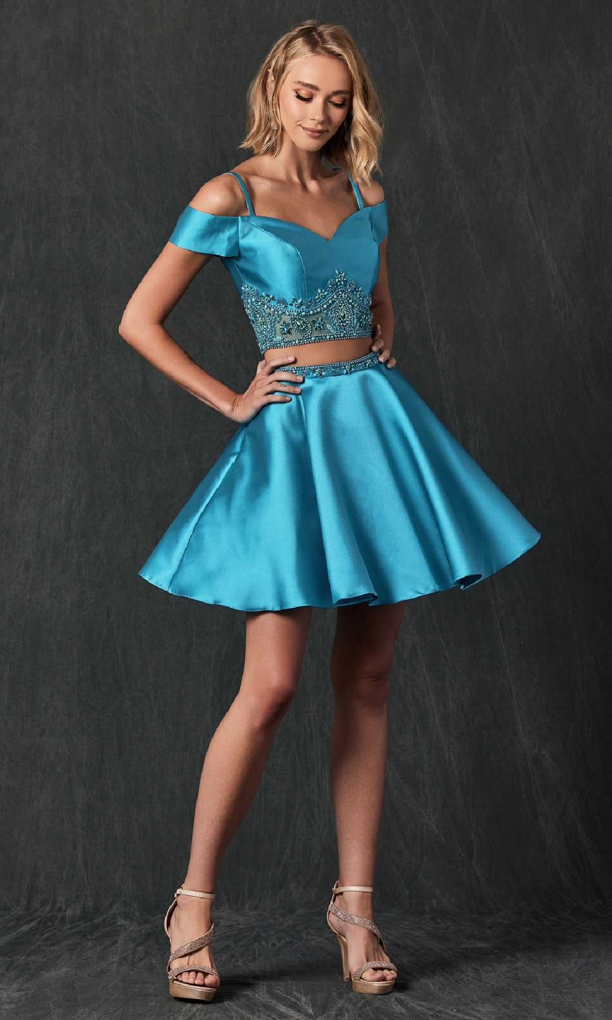 A woman models the Juliet 790, a short two-piece homecoming dress with an off-the-shoulder, bright blue embellished fitted bodice and flared A-line skirt, set against a dark textured background.